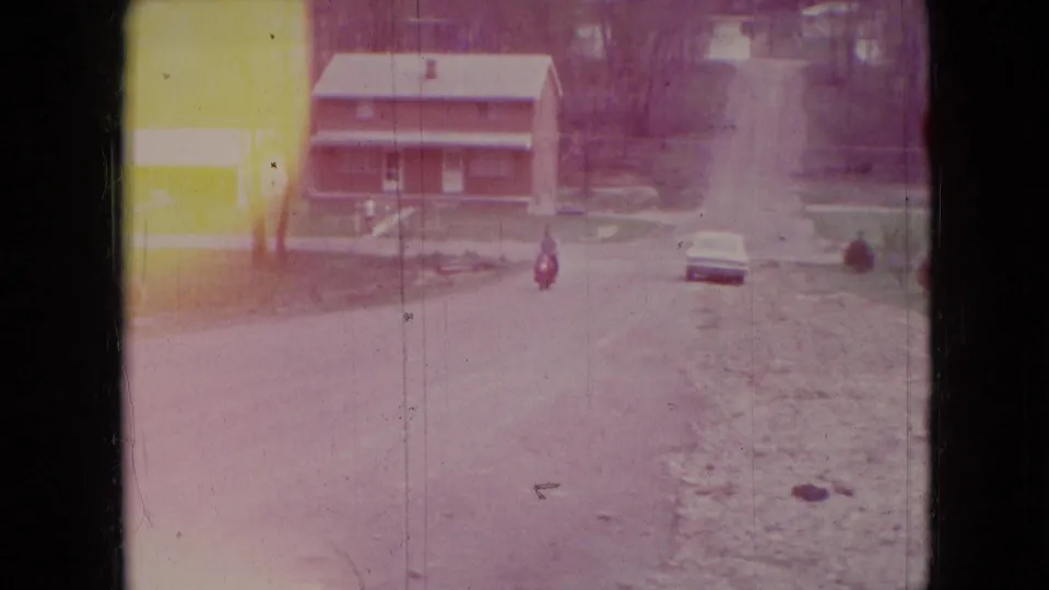 A residential street view in the supplied Athens, Ohio 1966 footage shows a brick house, a parked car, bare trees, and a small distant figure or object along the road.