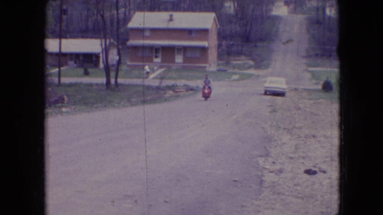 In supplied 1966 Athens, Ohio home-movie footage, a distant rider on a red motorcycle moves up a quiet residential road toward a two-story brick house with bare trees nearby.