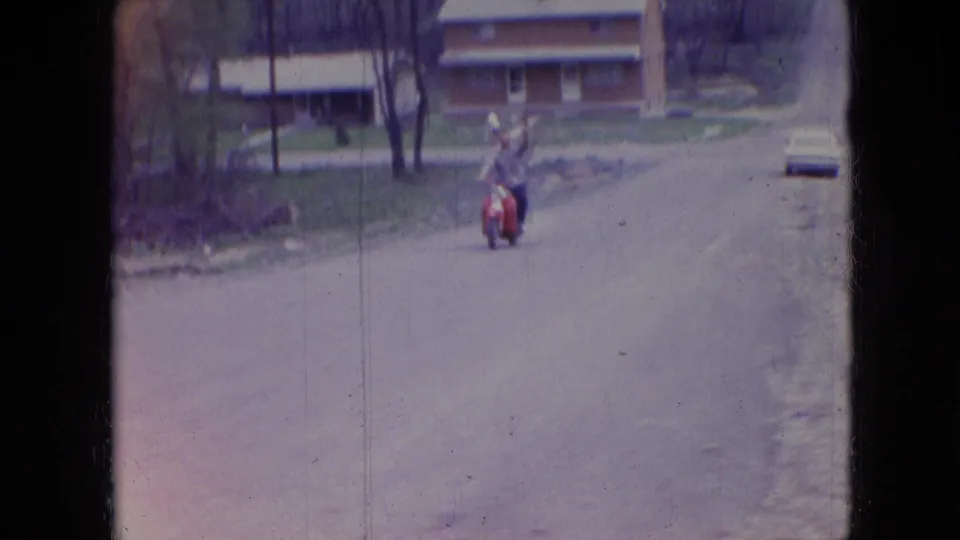 In the supplied Athens, Ohio 1966 clip, the red motorcycle rider is closer on the residential road, with a brick house, bare trees, and a parked light-colored car still visible.
