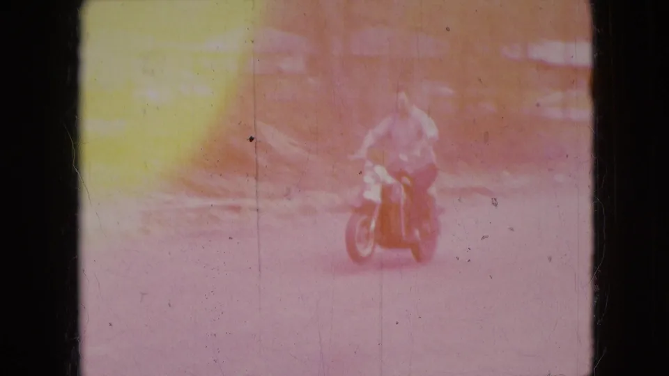 In 1966 home-movie footage from Athens, Ohio, a man on a red scooter approaches on a rural road through heavy color fading and film wear.