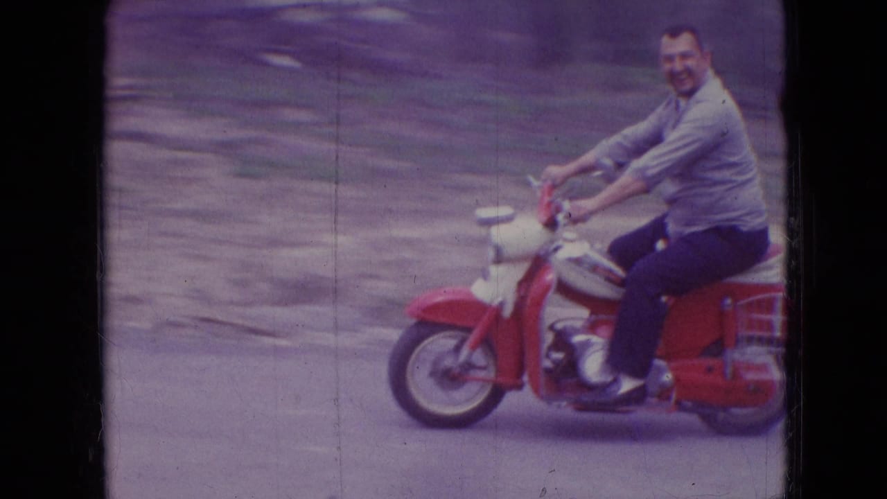 A smiling adult man in a light shirt and dark pants rides a red scooter past the camera on a rural road in the supplied 1966 Athens, Ohio footage.