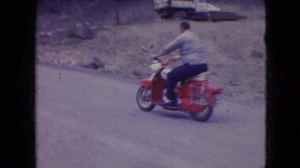 The rider continues along the rural road on a red scooter, with grassy banks and a parked car visible in the background of the 1966 Athens, Ohio home movie.