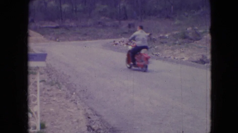 The man rides away on the red scooter along a curving countryside road, framed by trees, roadside grass, and faded 8mm film texture.