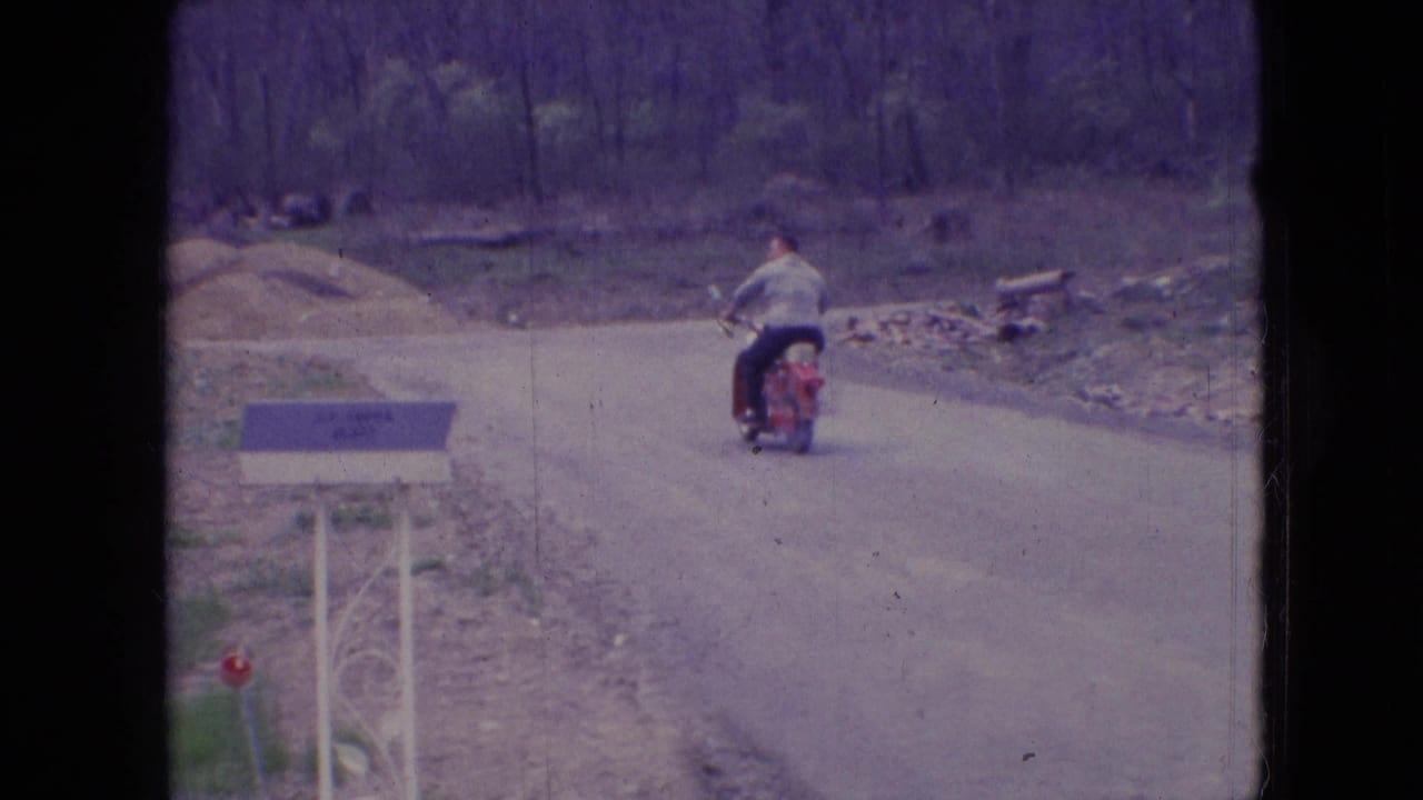In 1966 Athens, Ohio home-movie footage, a man in a light shirt rides a red motorcycle away along an unpaved road beside a decorative mailbox and wooded roadside.