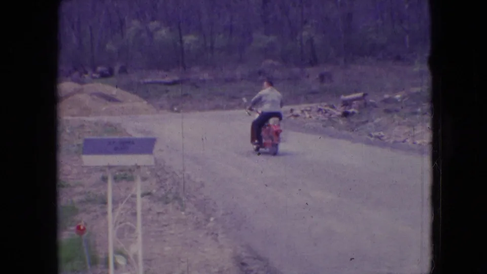 In 1966 Athens, Ohio home-movie footage, a man in a light shirt rides a red motorcycle away along an unpaved road beside a decorative mailbox and wooded roadside.