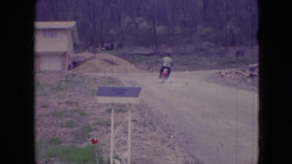 The rider on the red motorcycle recedes along the dusty road in Athens, Ohio, with trees, loose roadside material, and a small structure or construction area visible nearby.