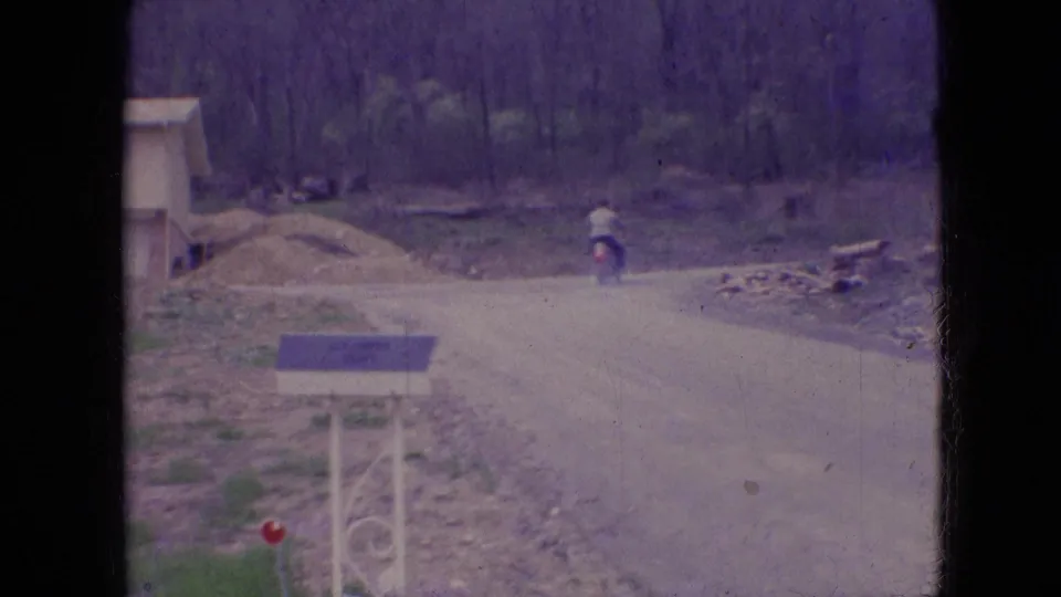 The motorcycle rider appears smaller in the frame as he travels away on a dusty rural road, with the mailbox foreground and wooded roadside still visible.