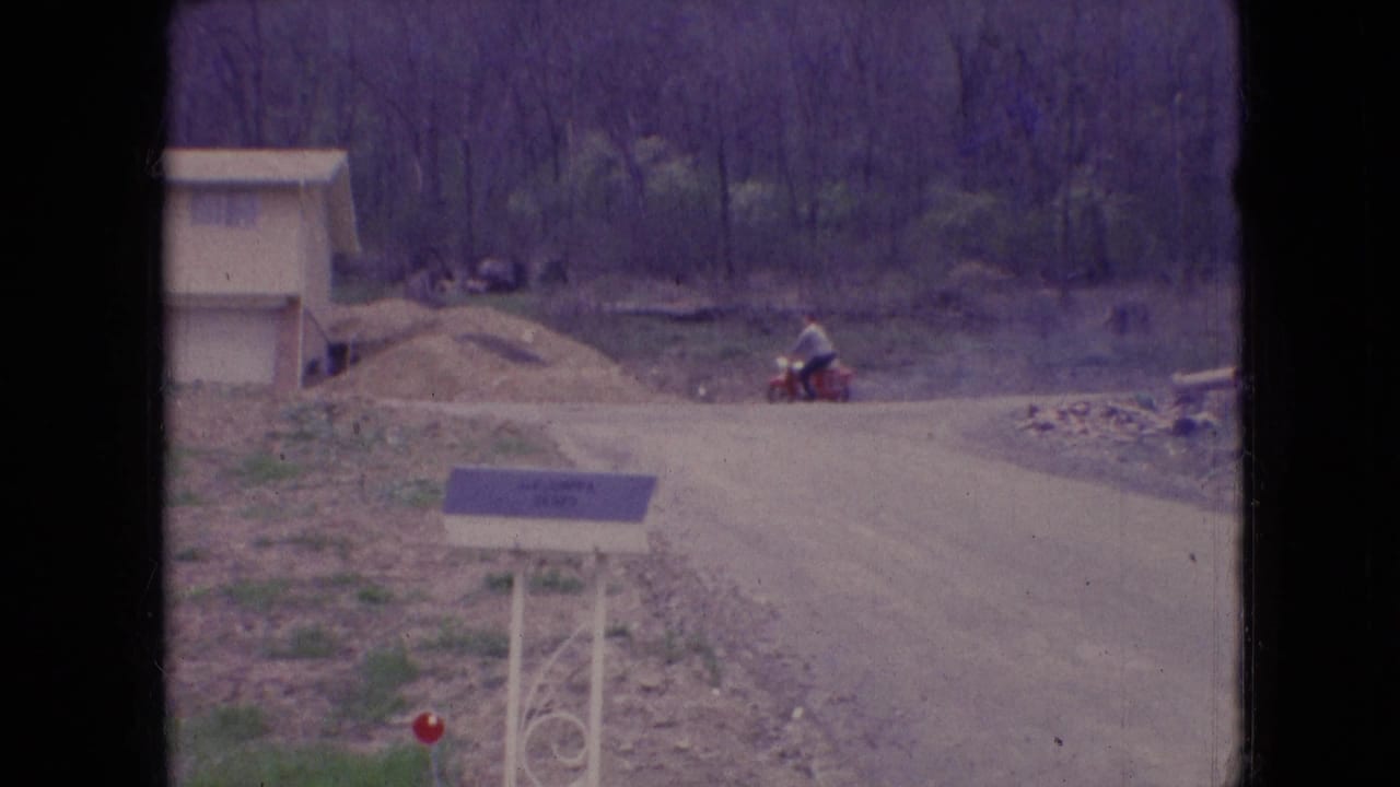 1966 Athens, Ohio 8mm frame showing a person on a red scooter moving along an unpaved residential road near a new house, graded soil, bare trees, and a foreground mailbox.