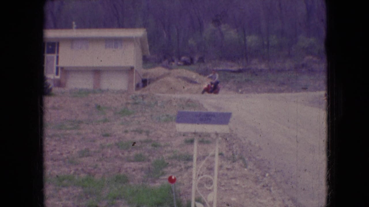 Red scooter rider farther along a dirt road beside a new house and open lot.