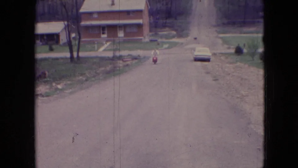 1966 Athens, Ohio 8mm frame looking down an unpaved road as a person rides a red scooter toward brick houses, a parked car, yards, and roadside construction debris.