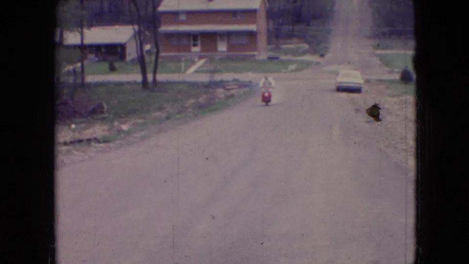 1966 Athens, Ohio 8mm frame showing a red scooter rider centered on a wide unpaved residential road, with brick houses, a pale parked car, bare trees, and rough roadside ground.