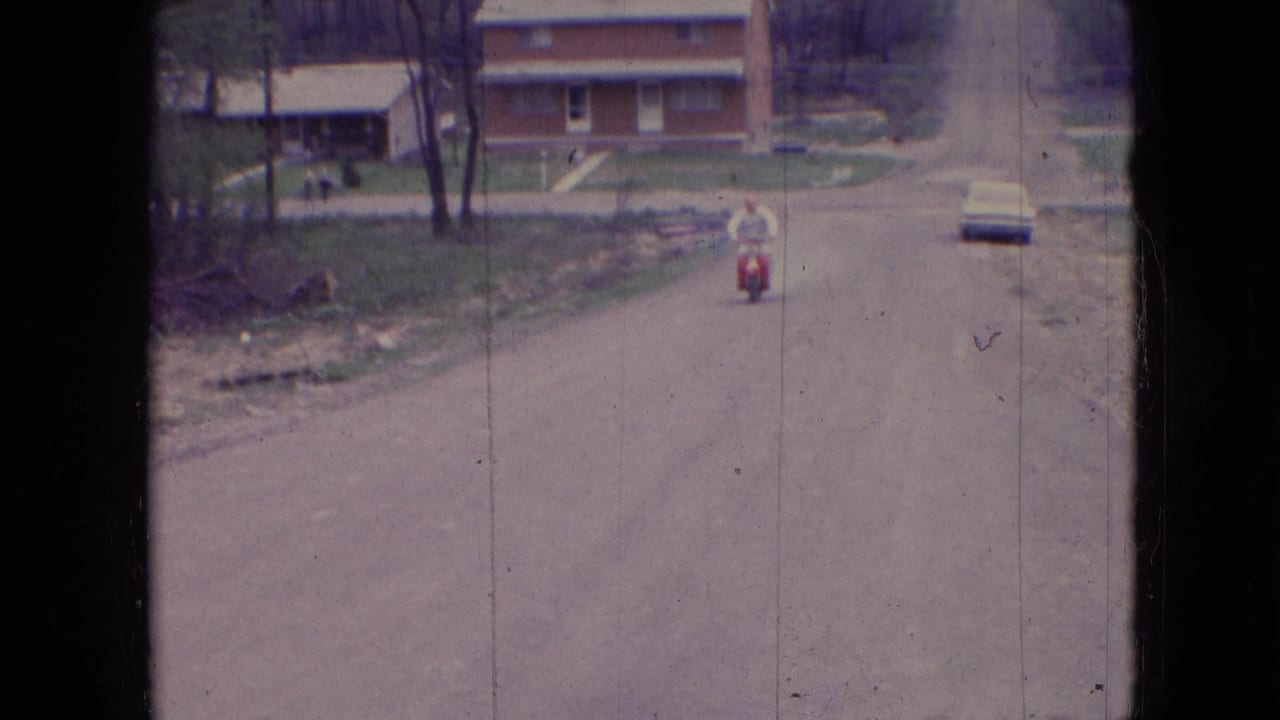 In Athens, Ohio in 1966, a distant adult man rides a red moped toward the camera along a quiet residential street with houses, trees, and a parked car beyond him.