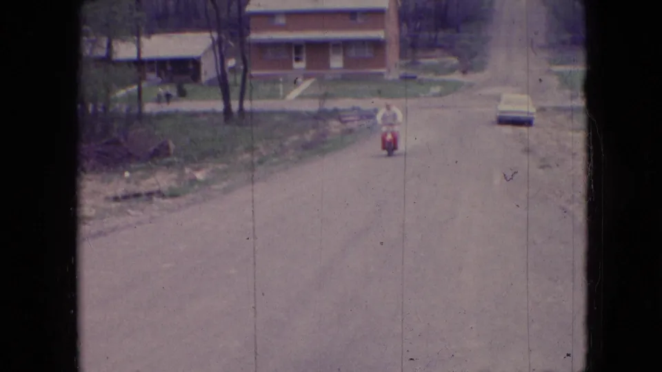 In Athens, Ohio in 1966, a distant adult man rides a red moped toward the camera along a quiet residential street with houses, trees, and a parked car beyond him.