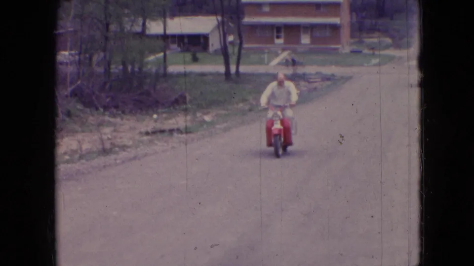 The bald adult rider smiles as he comes closer on a red moped, framed by a paved street, bare trees, lawns, and a brick house in the 1966 Athens, Ohio home-movie scene.