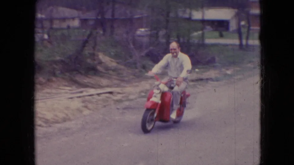 The rider passes closer to the camera on a red moped, smiling on a residential street in Athens, Ohio, with roadside dirt, trees, lawns, and houses behind him.