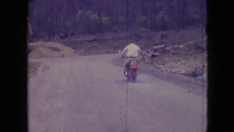 A distant view shows the man riding the red moped away on a quiet rural road, with trees and rough roadside ground visible in the background.
