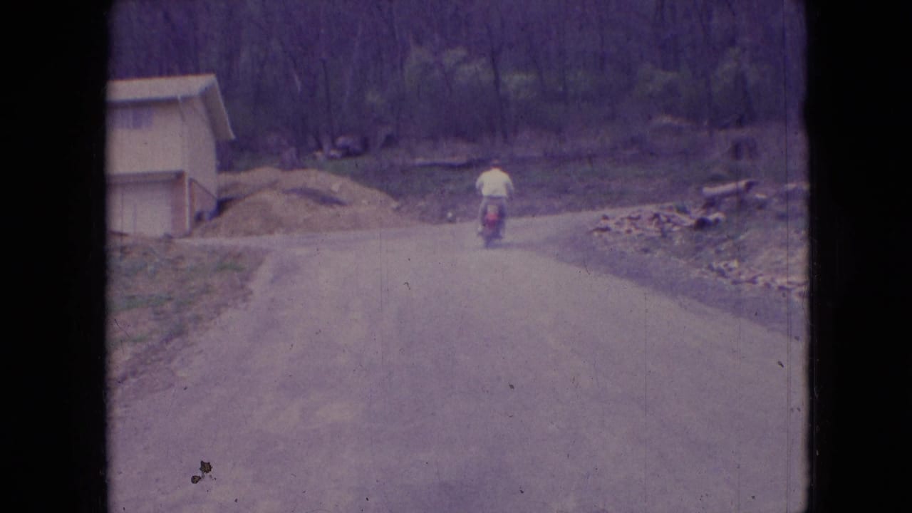 In 1966 Athens, Ohio home-movie footage, a man rides a small red scooter away from the camera along a rural dirt or gravel road bordered by woods and a small structure.