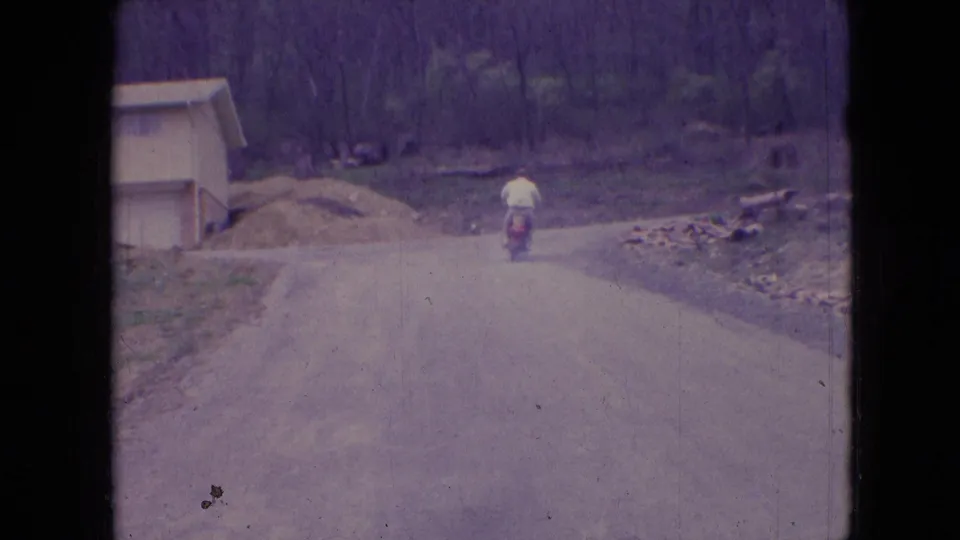 In 1966 Athens, Ohio home-movie footage, a man rides a small red scooter away from the camera along a rural dirt or gravel road bordered by woods and a small structure.