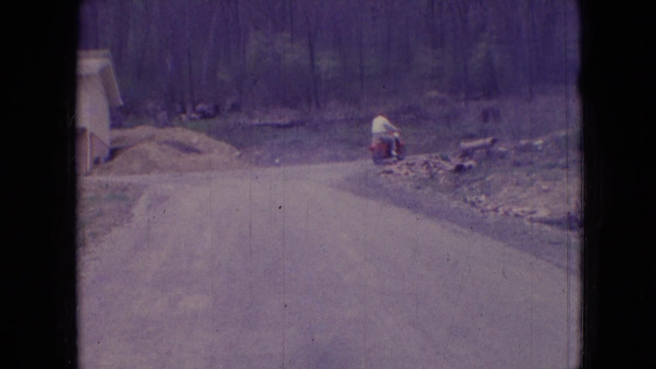 Scooter rider turns near the roadside bend with woods behind him.