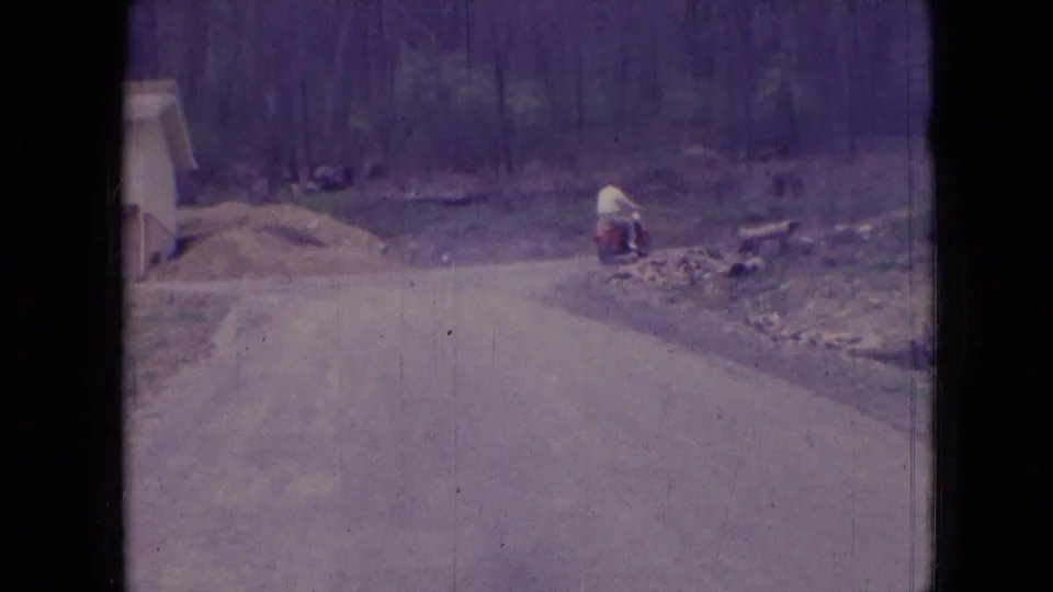 The man on the red scooter appears farther down the rural Athens, Ohio road, turning near a roadside bend with wooded ground and earth piles in the background.