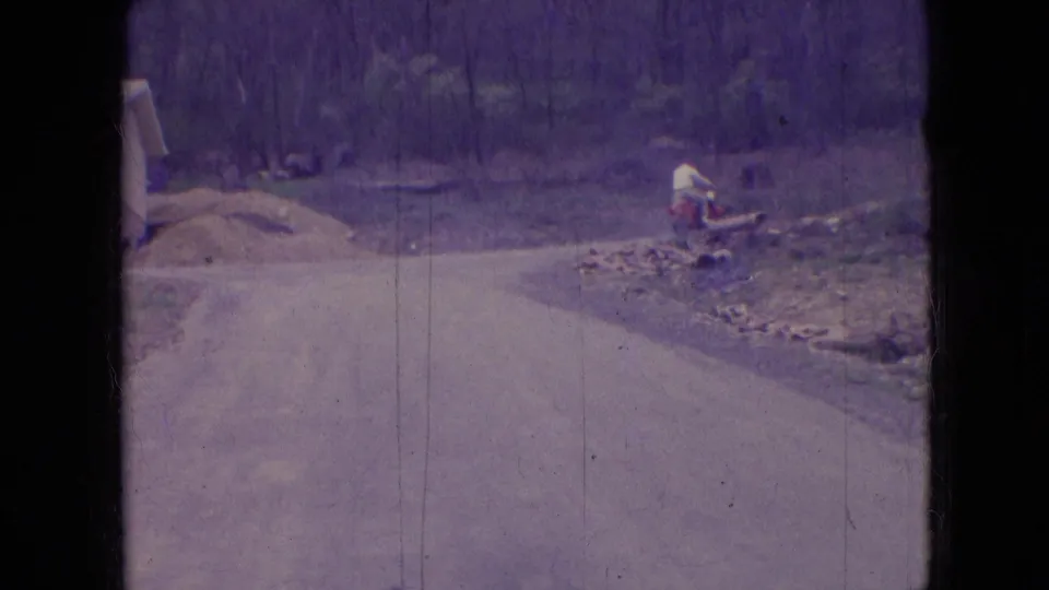 The scooter rider remains in the distance near the edge of a rural road, with a wooded slope, scattered logs or brush, and a small structure partly visible in the frame.