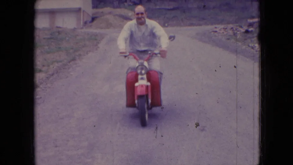 A bald adult man in a light long-sleeved shirt smiles while riding a red scooter toward the camera on a rural road in the 1966 Athens, Ohio home movie.