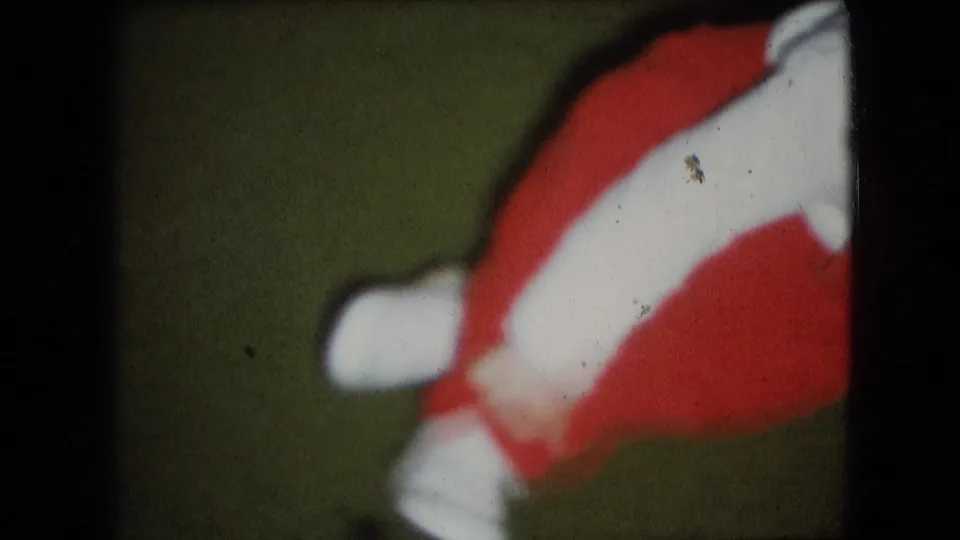 1966 Athens, Ohio 8mm frame showing the lower half of a small child in a red outfit with white straps and shoes, partly turned on a green carpet.
