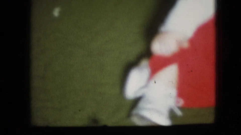 Close-cropped 1966 Athens, Ohio home-movie frame showing part of a small child in red clothing with a white shoe near the edge of a green floor.