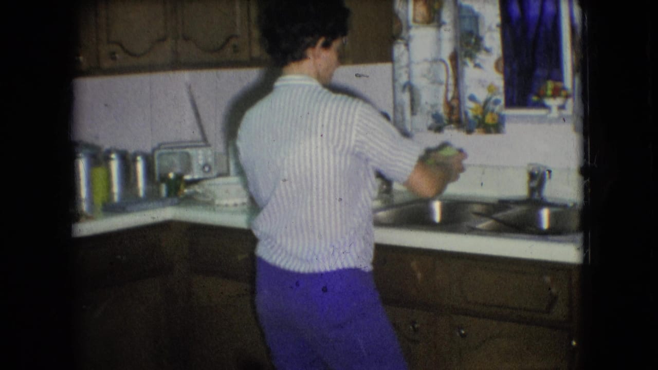 In 1966 Athens, Ohio home-movie footage, a woman in a striped shirt and purple pants stands at a double sink in a wood-cabinet kitchen.