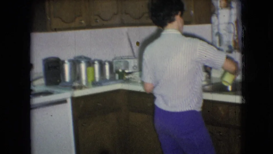The woman remains by the kitchen counter and sink in the Athens, Ohio home interior, with canisters, cabinets, and curtains visible around her.