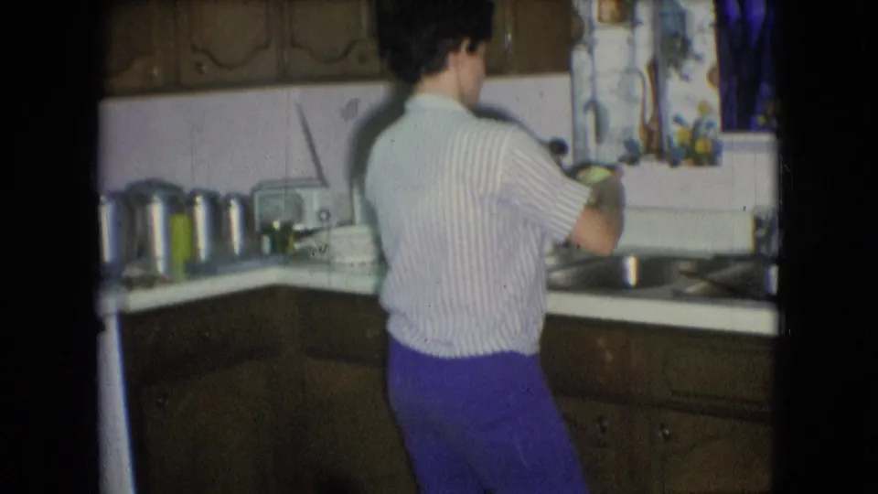 A woman holds a green sponge-like object near the double sink in a 1966 Athens, Ohio kitchen, framed by cabinets, curtains, and countertop items.