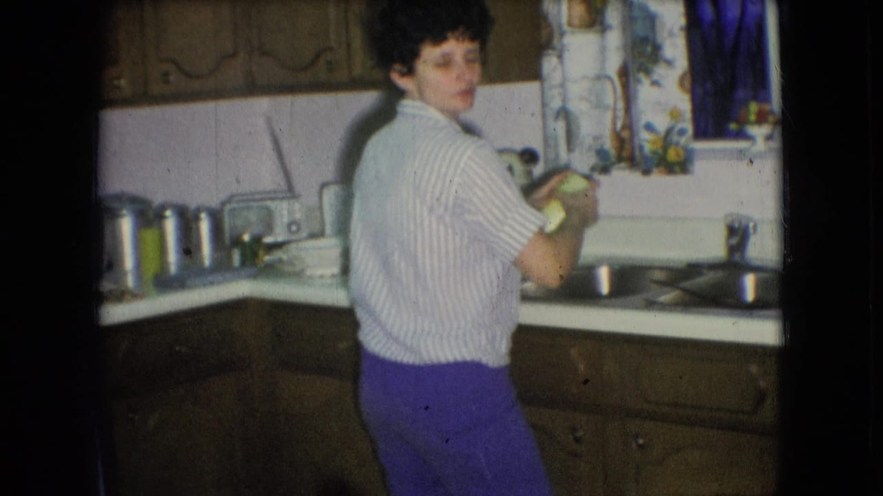 A young person in a striped shirt and purple trousers stands at a sink in an Athens, Ohio kitchen, 1966, with wood cabinets, white backsplash, and a patterned curtain visible.