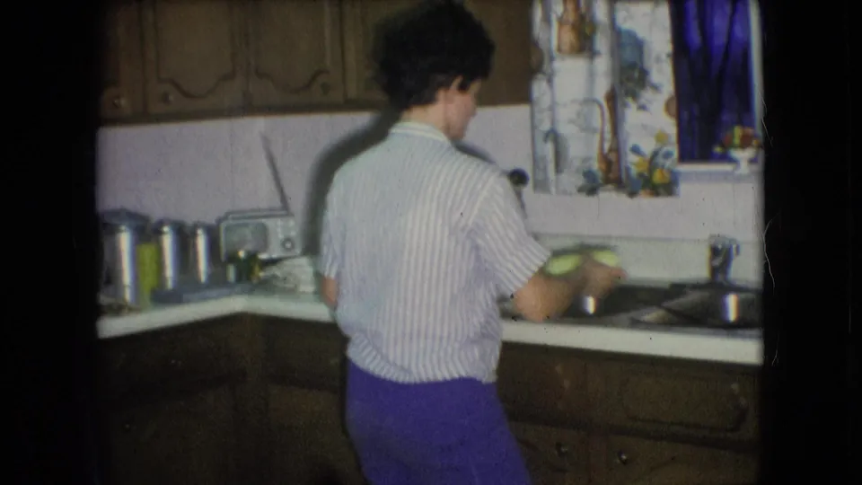 The person faces the sink in the 1966 Athens, Ohio kitchen as dishes are handled near the metal basin, with cabinets and a curtained window area in view.