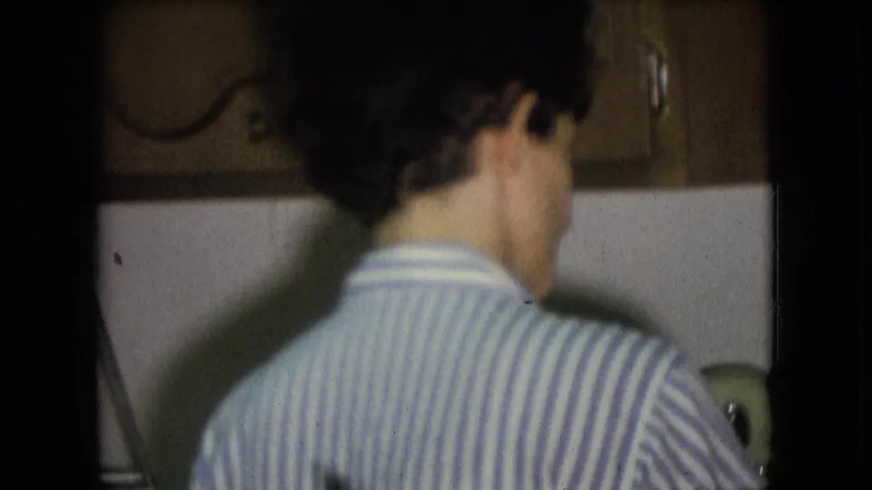 The final frame stays close behind the person in the striped shirt as they continue dishwashing beside wood cabinets and a white backsplash in the 1966 home interior.