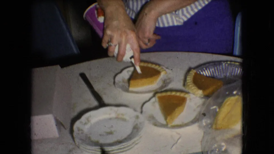 The woman works over a slice of sweet potato pie at the kitchen table, with stacked plates, a knife, pie tins, and other dessert portions visible in the home interior.