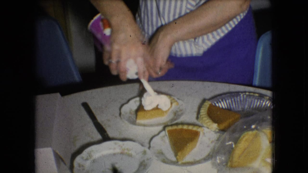 In a 1966 Athens, Ohio home kitchen, hands top a pumpkin pie slice with whipped cream beside plates and other pie slices on a white table.