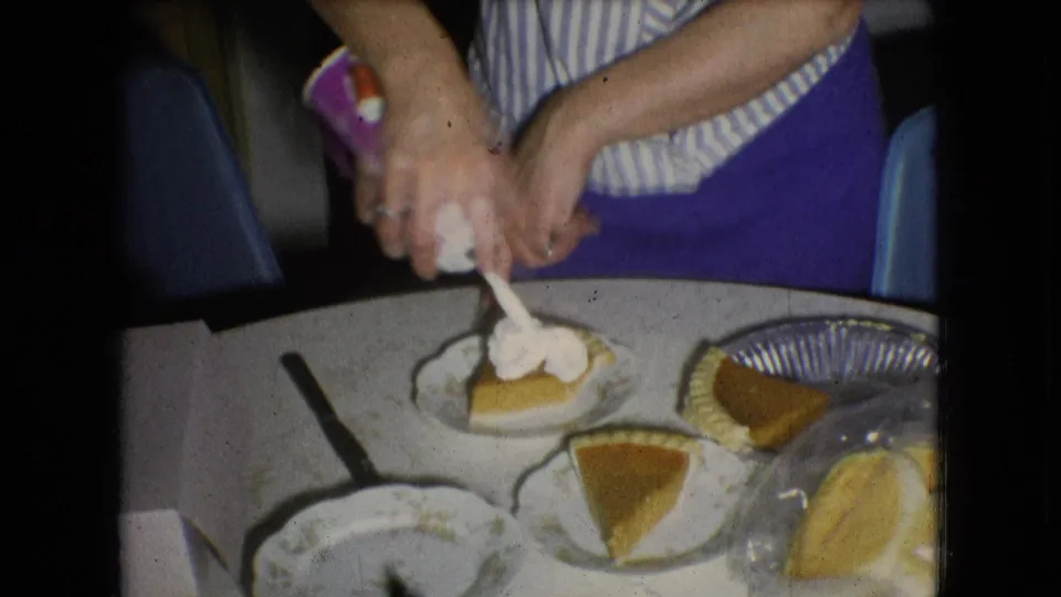In a 1966 Athens, Ohio home kitchen, hands top a pumpkin pie slice with whipped cream beside plates and other pie slices on a white table.