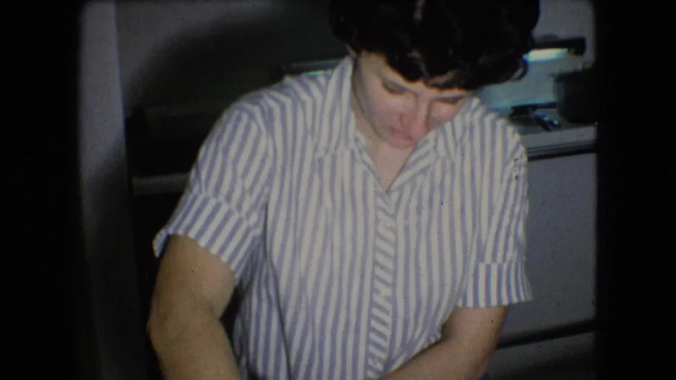A woman in a striped shirt leans over a table in the Athens, Ohio home kitchen, with a small appliance and domestic interior visible behind her.