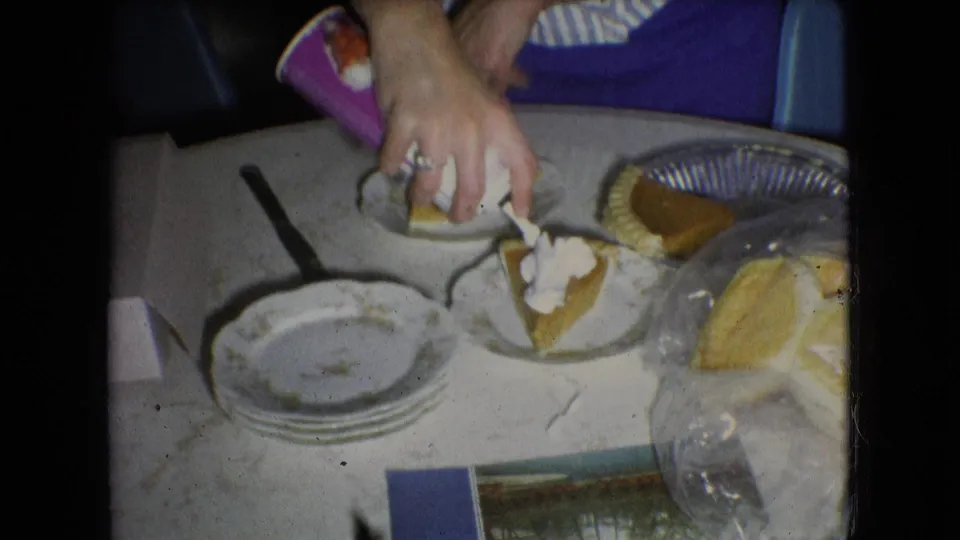 Hands top pumpkin pie slices with whipped cream on a white kitchen table in 1966 Athens, Ohio, with plates, a pie tin, and serving items nearby.