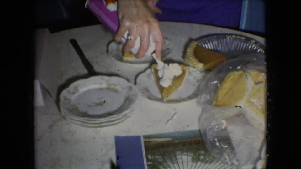 In 1966 Athens, Ohio home-movie footage, a woman's hands place whipped cream on pumpkin pie slices beside stacked plates, a whole pie, and wrapped bread on a kitchen table.