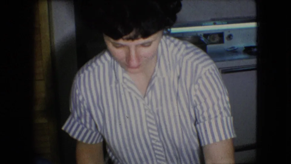 A woman in a striped blouse looks downward in a private Athens, Ohio kitchen, with soft 8mm focus and kitchen appliances visible in the background.