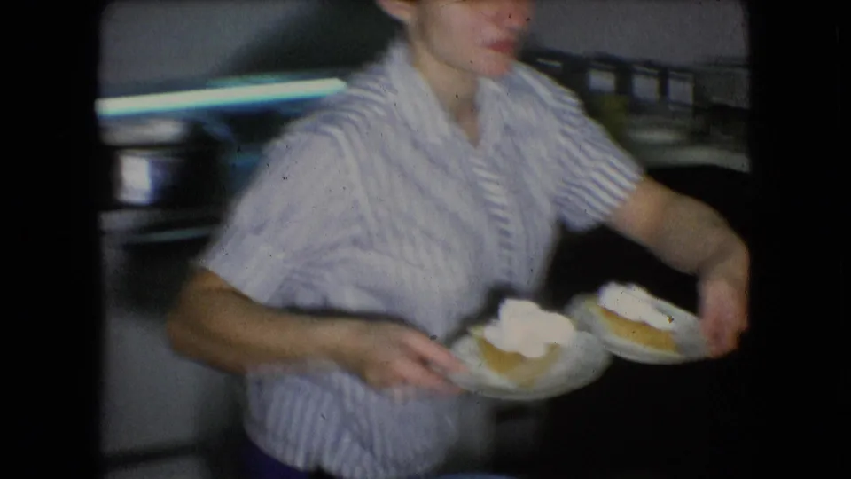 The woman in a striped blouse carries paper plates of pumpkin pie with whipped cream through the private kitchen in this 1966 Athens, Ohio home movie.