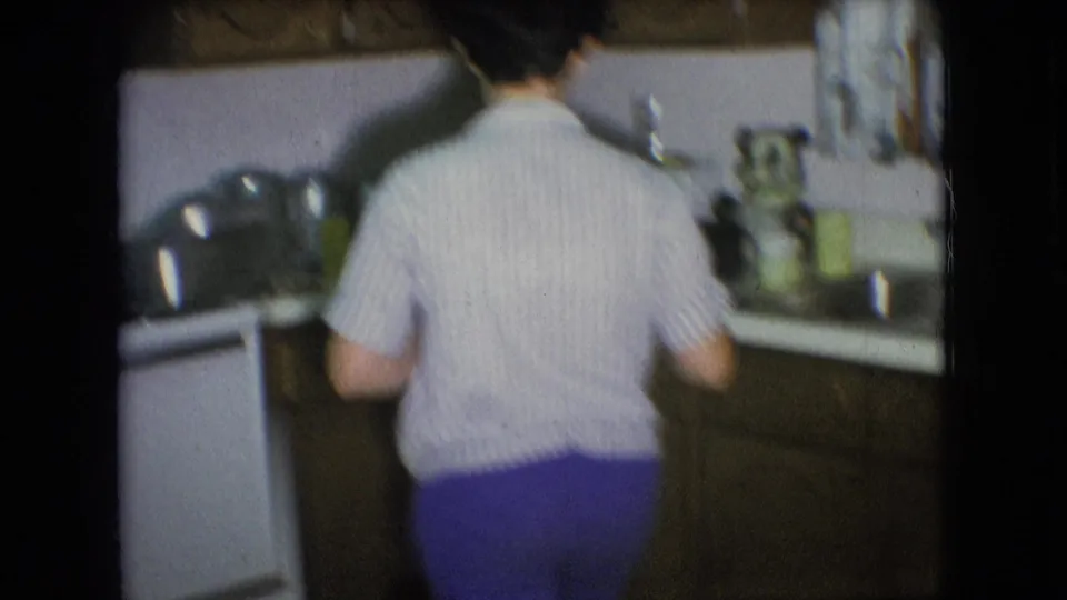In the 1966 Athens, Ohio home-movie footage, an adult is seen from behind at a kitchen counter in a softly focused private interior.