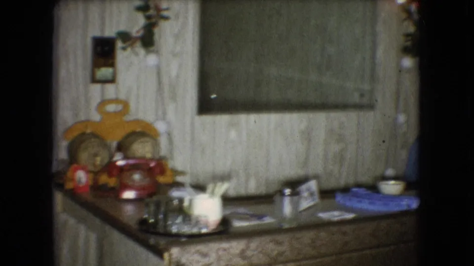 Countertop area with a red rotary phone, glassware, small holiday decorations, a wall-hung board or mirror, and wood paneling in a 1966 Athens, Ohio home movie frame.