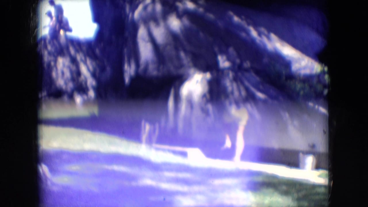 A woman stands or walks near grass below tall rock formations in the wooded Athens, Ohio setting, seen through soft, color-shifted 8mm footage.