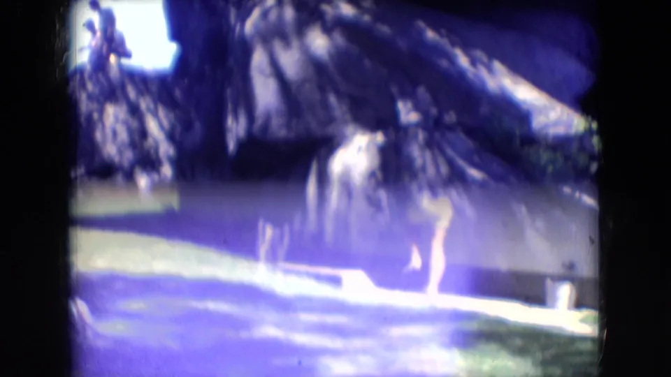 A woman stands or walks near grass below tall rock formations in the wooded Athens, Ohio setting, seen through soft, color-shifted 8mm footage.