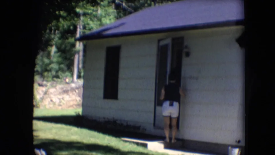 A woman in a dark top and white shorts approaches a small light-colored cabin with a dark roof in the wooded Athens, Ohio home-movie scene.