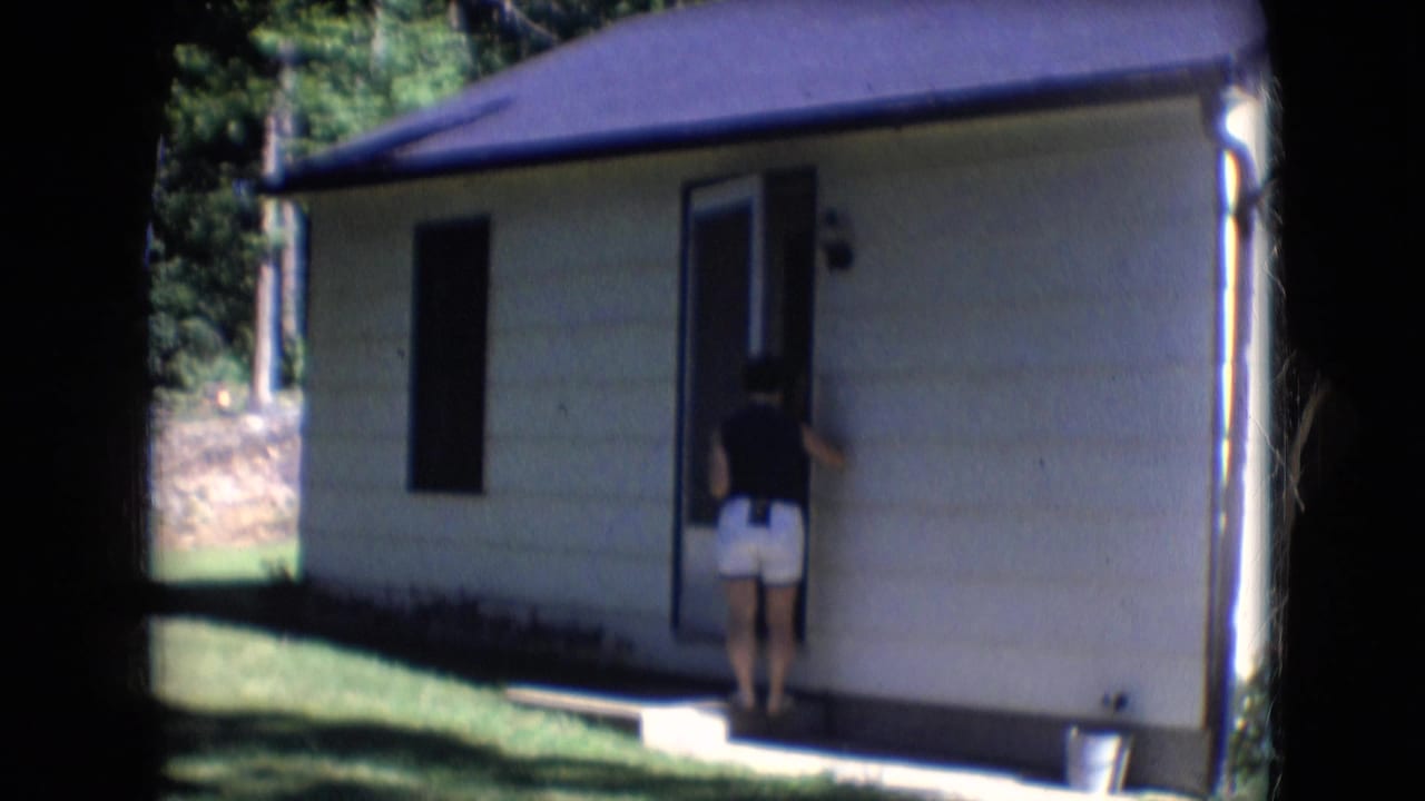 1966 Athens, Ohio 8mm frame showing a woman in a dark top and white shorts standing at an open door beside a lawn and trees.