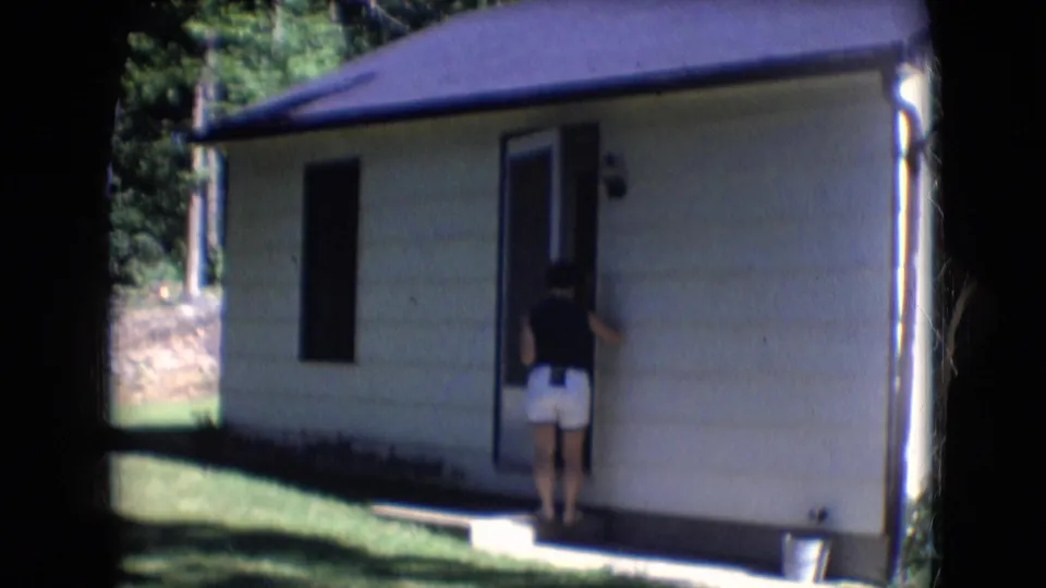 1966 Athens, Ohio 8mm frame showing a woman in a dark top and white shorts standing at an open door beside a lawn and trees.