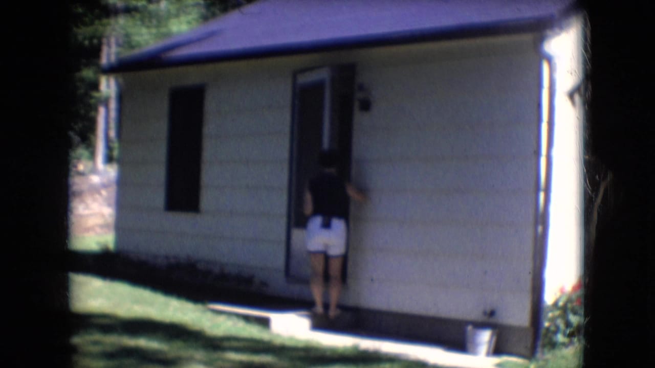 1966 Athens, Ohio home-movie frame with a woman seen from behind at the open door of a residential house, with lawn and trees around it.
